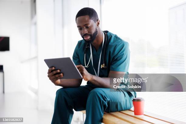 male doctor sitting in hospital hallway, looking at digital tablet - healthcare facilities stock pictures, royalty-free photos & images