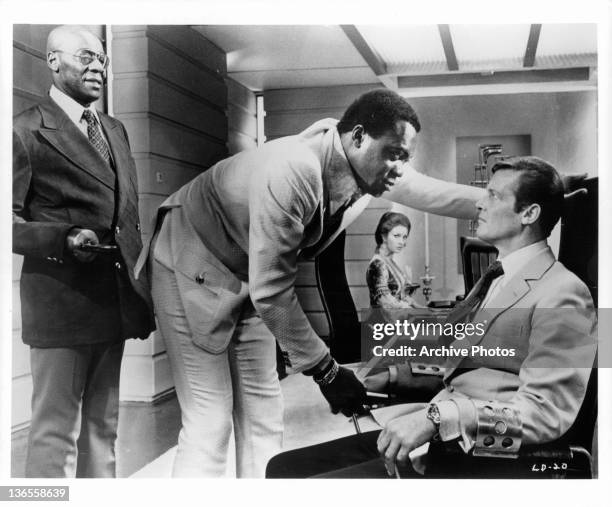 Roger Moore sitting in a chair with arm restraints while Yaphet Kotto stares at him closely face to face in a scene from the film 'Live And Let Die',...