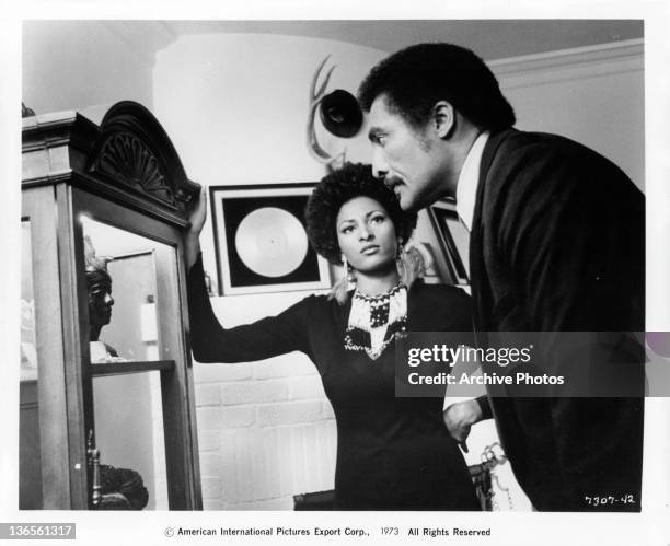 Pam Grier resting her hand on a wooden display case while William Marshall is staring mesmerized of what he sees inside the case in a scene from the...