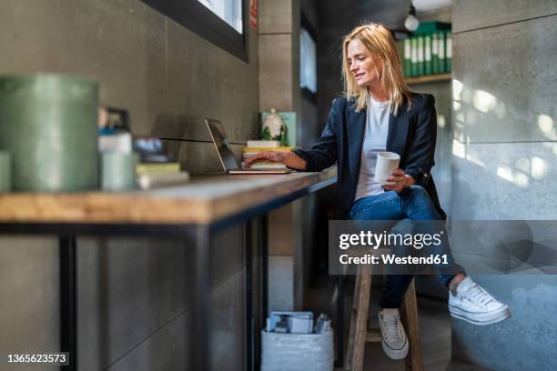 businesswoman with coffee mug using laptop sitting on stool at office - benen over elkaar geslagen stockfoto's en -beelden