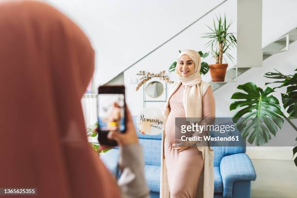 woman photographing pregnant friend on mobile phone in living room - pregnant muslim stock pictures, royalty-free photos & images