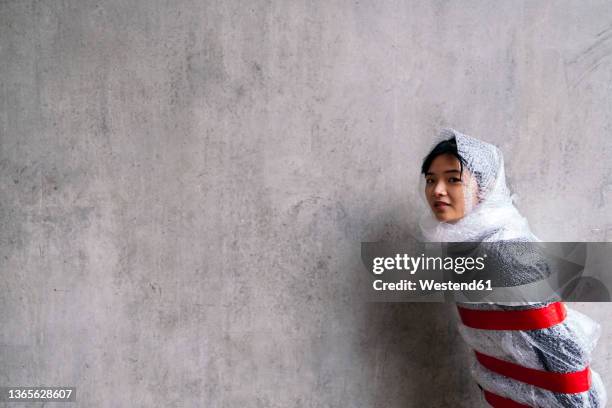 young woman protected in bubble pack by gray wall - bubble wrap stock pictures, royalty-free photos & images