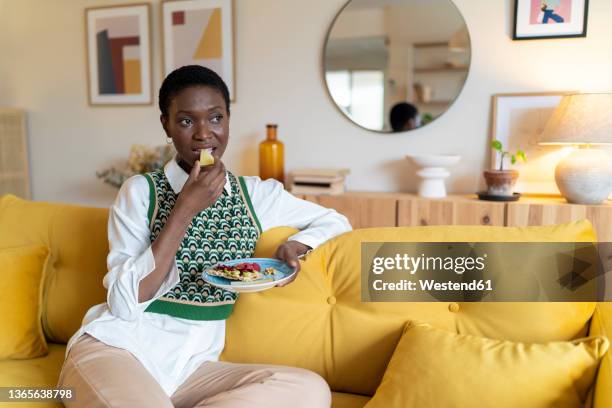 thoughtful woman eating fruits on sofa at home - snack photos et images de collection