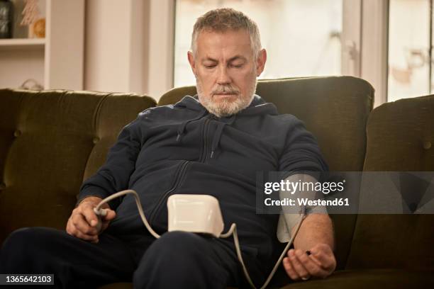 senior man checking blood pressure sitting on sofa at home - atherosclerosis stock pictures, royalty-free photos & images