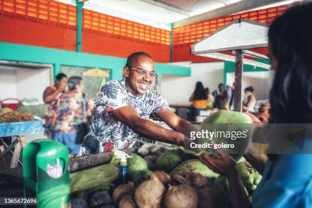 smiling young market vendor giving fresh coconut to female client - feirante imagens e fotografias de stock