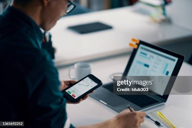 mid adult man checking financial information on a smart phone while doing his bookkeeping - smart numbers office stockfoto's en -beelden