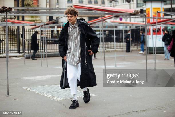 Actor Maxence Danet-Fauvel wears a black jacket, gray striped button up shirt, oversized black pants, white socks, and black shoes at the AMI Paris...