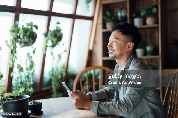 portrait of smiling young asian man with smartphone sitting at home by the window with green plants, relaxing and having a quiet moment - sustainability asia stock pictures, royalty-free photos & images