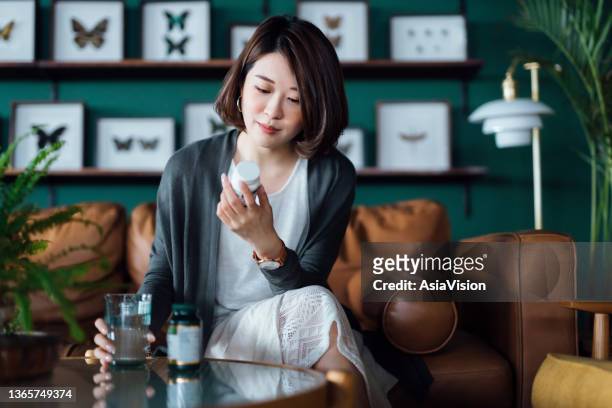 junge asiatische frau, die medikamente mit einem glas wasser auf dem couchtisch einnimmt und die informationen auf dem etikett ihrer medikamente zu hause liest. gesundheitskonzept - qual der wahl stock-fotos und bilder