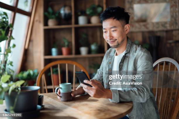 smiling young asian man using smartphone and having coffee in the balcony at home. the use of technology in everyday life - asian using phone stock pictures, royalty-free photos & images