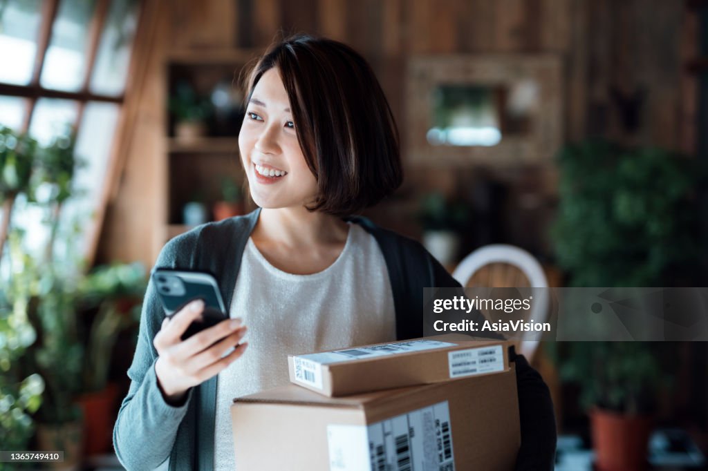 Schöne lächelnde junge Asiatin mit Smartphone, die Pakete mit Hauslieferdienst zu Hause empfängt. Online-Shopping, Mobile Payment. Angenehmes Einkaufserlebnis