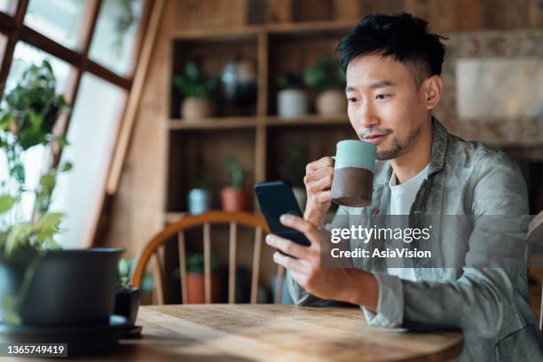 junger asiatischer mann, der online-banking mit mobiler app auf dem smartphone verwaltet, sich um sein geld und seine finanzen kümmert, während er sich zu hause entspannt. banking mit technologie - cafehaus stock-fotos und bilder