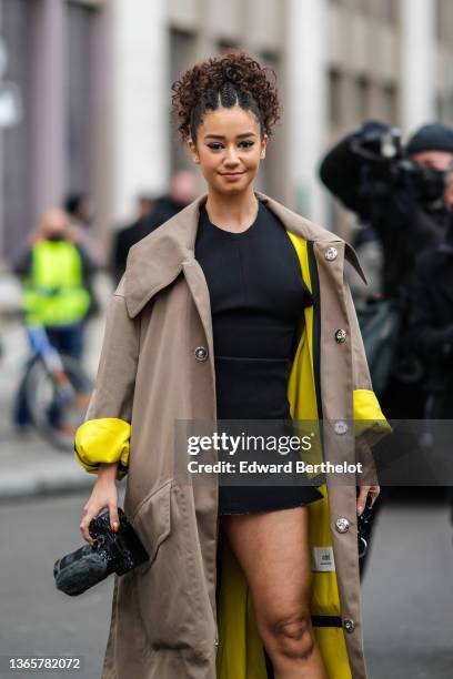 Lena Mahfouf aka Lena Situations wears a black tube short slit / split dress, a beige with yellow interior long trench coat from Ami, a black shiny...