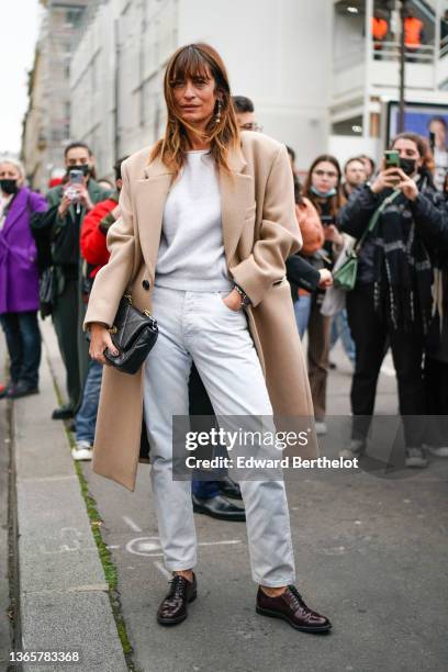 Caroline de Maigret wears a gold pendant earring, a white cashmere pullover, a beige wool long coat, white denim jeans pants, a black shiny leather...