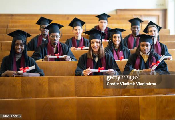 happy multiethnic group of students sitting in the university lecture hall, holding their diplomas - auditorium stock pictures, royalty-free photos & images