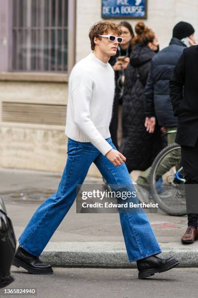 Guest wears a white wool pullover, high waist blue denim flared jeans pants, white sunglasses, black shiny leather ankle boots, outside AMI, during...