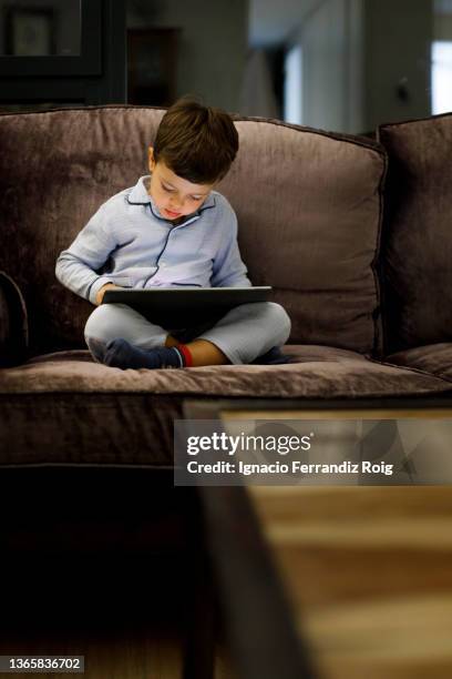 handsome little boy in pijama sitting with an electronic tablet on the sofa at home before going to sleep. - relajación photos et images de collection