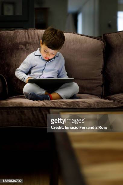 handsome little boy in pijama sitting with an electronic tablet on the sofa at home before going to sleep. - relajación - fotografias e filmes do acervo