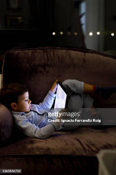 handsome boy in pijama playing with electronic tablet lying on the sofa at home. - tecnología foto e immagini stock
