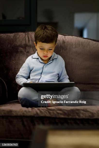 handsome little boy in pijama sitting with an electronic tablet on the sofa at home before going to sleep. - relajación - fotografias e filmes do acervo