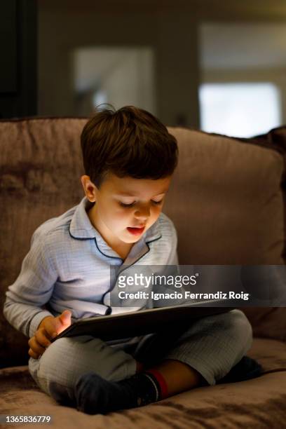 handsome little boy in pijama sitting with an electronic tablet on the sofa at home before going to sleep. - niñez stock-fotos und bilder