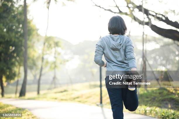 stretching may decrease your risk of injury during workout. rear view of asian female runner doing quadriceps standing before morning jogging on trails in city park. - standing on one leg fotografías e imágenes de stock