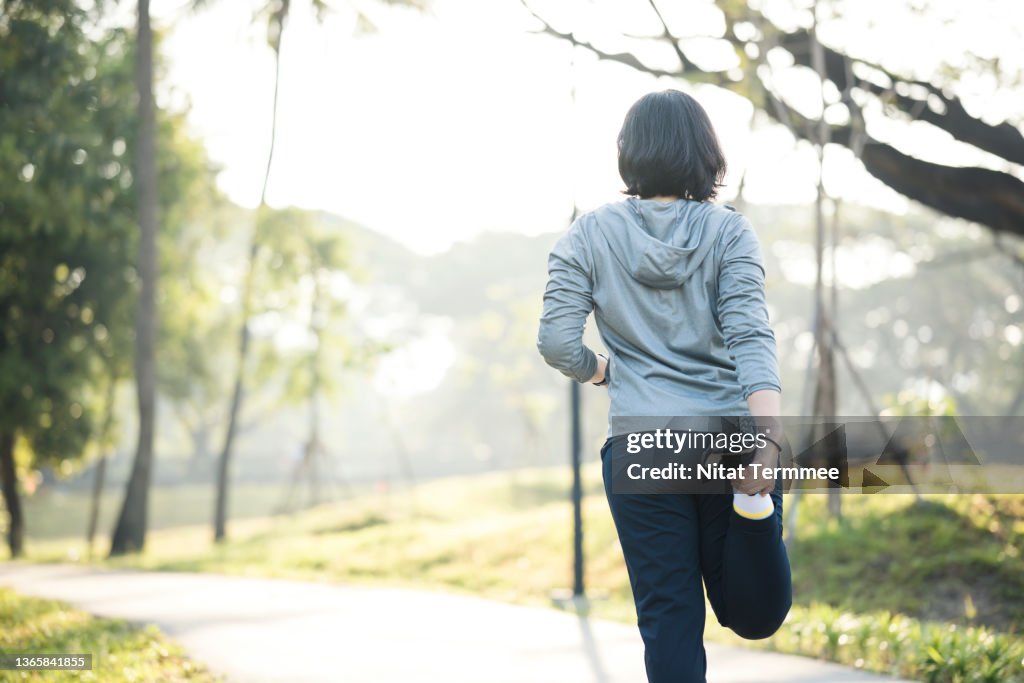 Stretching may decrease your risk of injury during workout. Rear View of Asian female runner doing quadriceps standing before morning jogging on trails in city park.