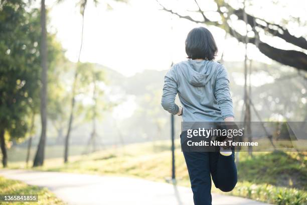 stretching may decrease your risk of injury during workout. rear view of asian female runner doing quadriceps standing before morning jogging on trails in city park. - woman standing exercise stockfoto's en -beelden