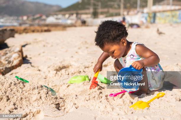 little girl playing with a sand pail and shovel at the beach - african girls on beach stock pictures, royalty-free photos & images