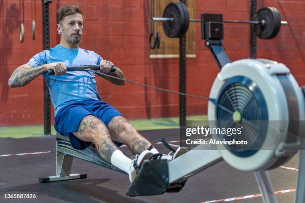 tattooed man rowing on ergometer in gym crosstraining box wearing blue sportswear - aviron photos et images de collection