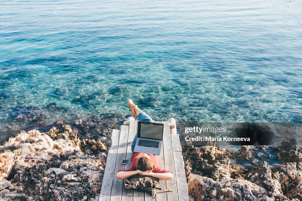 Young freelance woman works in laptop lying at backpack on wooden beach decks against Mediterranean sea