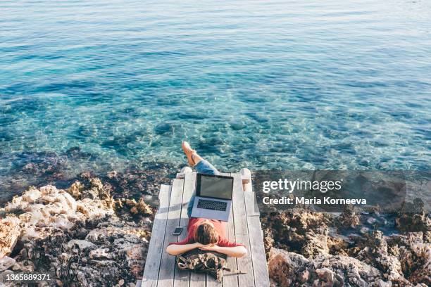 young freelance woman works in laptop lying at backpack on wooden beach decks against mediterranean sea - remote location fotografías e imágenes de stock