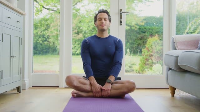 Portrait Of Smiling Mature Man Sitting On Mat At Home In Yoga Position Meditating