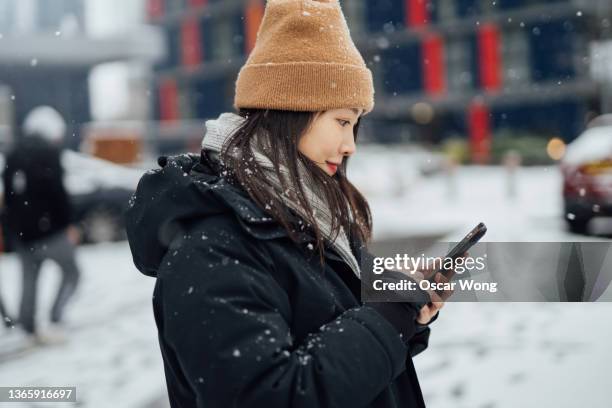 smiling young asian woman in warm clothes using smartphone while standing in snow city - new york city snow stock pictures, royalty-free photos & images