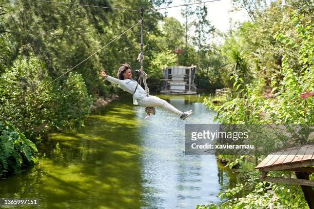 smiling young black woman riding zip line across pond - ziplining stock pictures, royalty-free photos & images