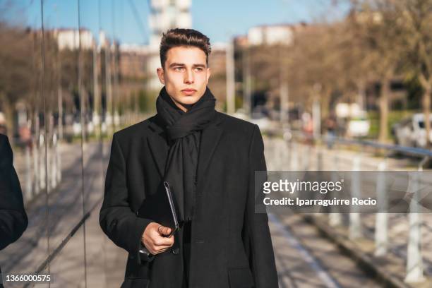 portrait of a fashionable young caucasian businessman outside the office in winter, long trench coat and black scarf, with briefcase and tablet heading to work walking - heading design stock pictures, royalty-free photos & images