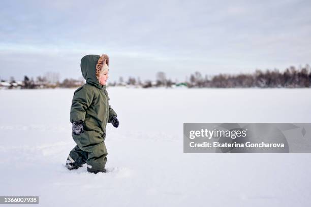 a 3-year-old boy in a green winter jumpsuit walks through the snow in nature. - snow angel stock pictures, royalty-free photos & images
