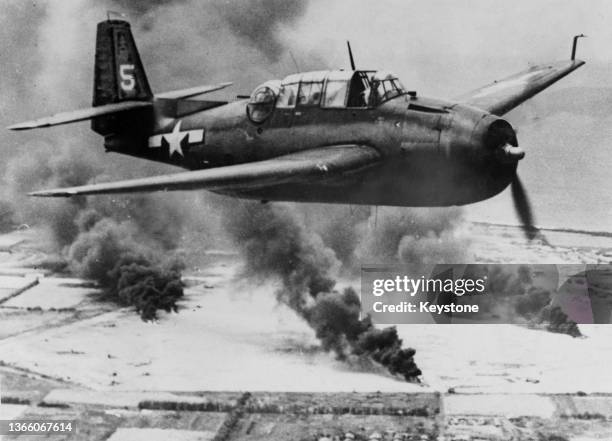 United States Navy Grumman TBF-1C Avenger carrier-based torpedo bomber surveys damaged aircraft and instillations burning following an attack on the...