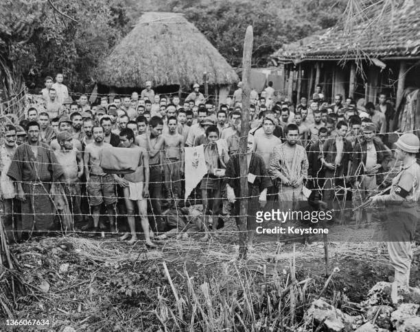 Crowd of Japanese prisoners of war from the Imperial Japanese Army's Thirty-Second Army stand behind a barbed wire fence, inside a stockade camp...