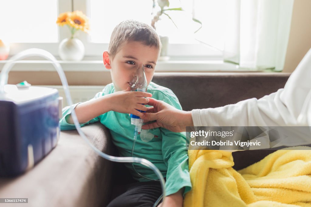 Sad sick boy breathing into inhalation mask his mom is helping him hold