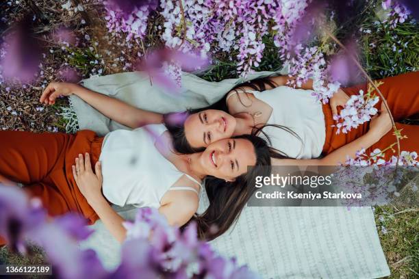 two young european twins with long dark hair lying on a picnic looking at the camera smiling faces close to each other top view through wisteria flowers - women dressed the same stock pictures, royalty-free photos & images