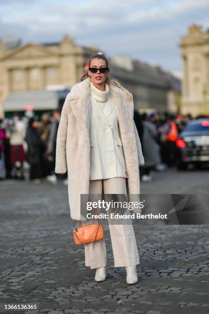 Guest wears sunglasses, a white wool turtleneck pullover, a white fluffy long winter coat, an orange leather crocodile pattern bag, a white double...