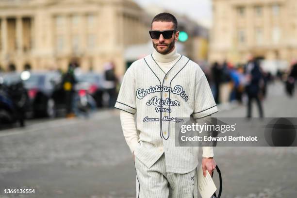 Pelayo Diaz wears sunglasses, a white wool turtleneck ribbed pullover, a white base ball oversized striped t-shirt with printed "Christian Dior...