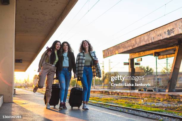 three beautiful young female friends walking at a train station with two suitcases during sunset while waiting to travel by train - holiday travel ahead of thanksgiving clogs airports highways and train stations stockfoto's en -beelden