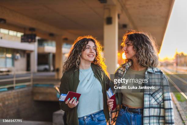zwei wunderschöne lockige freundinnen lächeln und unterhalten sich an einem bahnhof, während sie bei sonnenuntergang ihre pässe und tickets in der hand halten - vacances plage stock-fotos und bilder