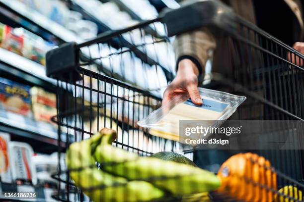 close up of woman pushing a shopping trolley, grocery shopping in supermarket. she is putting a packet of sliced cheese into the shopping cart. routine grocery shopping. healthy eating lifestyle - refrigerated section supermarket stock pictures, royalty-free photos & images