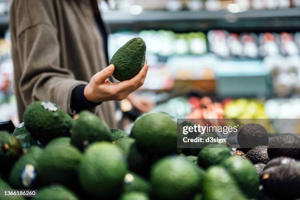 cropped shot of young asian woman grocery shopping for fresh organic fruits and vegetables in supermarket, close up of her hand choosing avocados along the produce aisle. routine grocery shopping. healthy eating lifestyle - ripe avocado fotografías e imágenes de stock