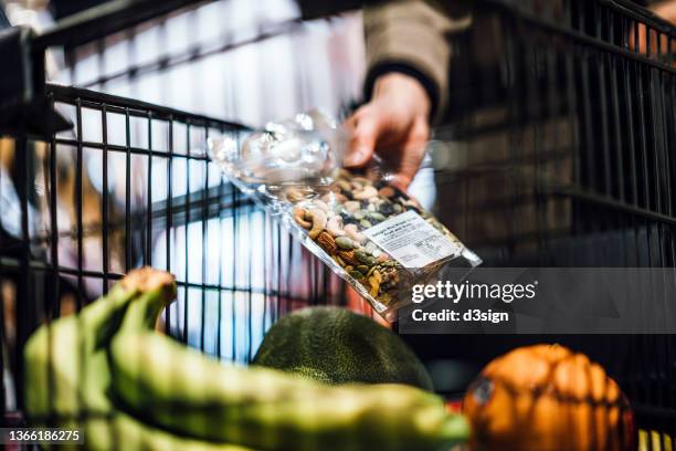 close up of woman pushing a shopping trolley, grocery shopping in supermarket. she is putting a packet of mixed nuts into the shopping cart. routine grocery shopping. healthy eating lifestyle - keto stock pictures, royalty-free photos & images