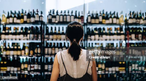 rear view of young asian woman grocery shopping for wines in a supermarket. she is standing in front of the liquor aisle and have no idea which wine to choose from - alcohol and women stock-fotos und bilder