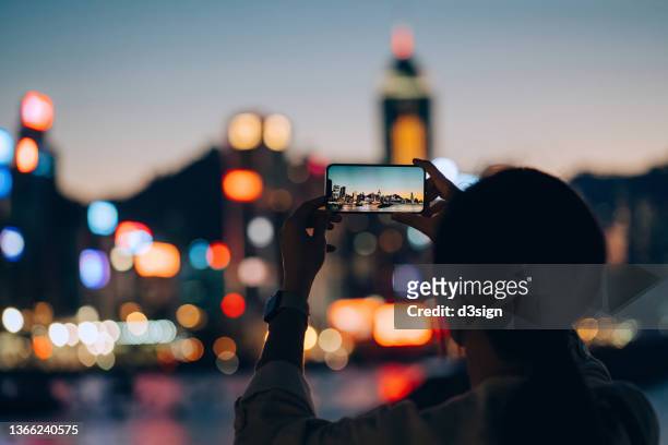 silhouette of young asian woman standing by the promenade, photographing hong kong city skyline at sunset with smartphone, with the view of multi-coloured and illuminated bokeh of the city. travel and city exploration concept - hometown stock-fotos und bilder